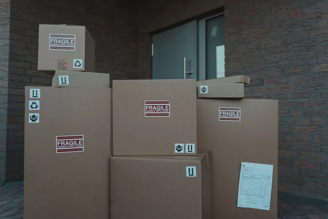 Several cardboard packages marked as fragile stacked at a building entrance. Versandkosten Ausland kalkulieren: Gewicht, Zone, Zuschläge